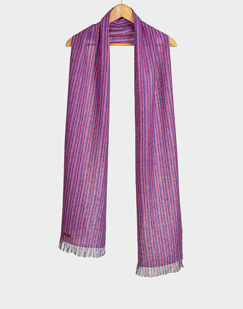 Chalina con rayas verticales en tonos púrpura fucsia y azul con flecos cortos colgada en gancho de madera diseño vibrante artesanal